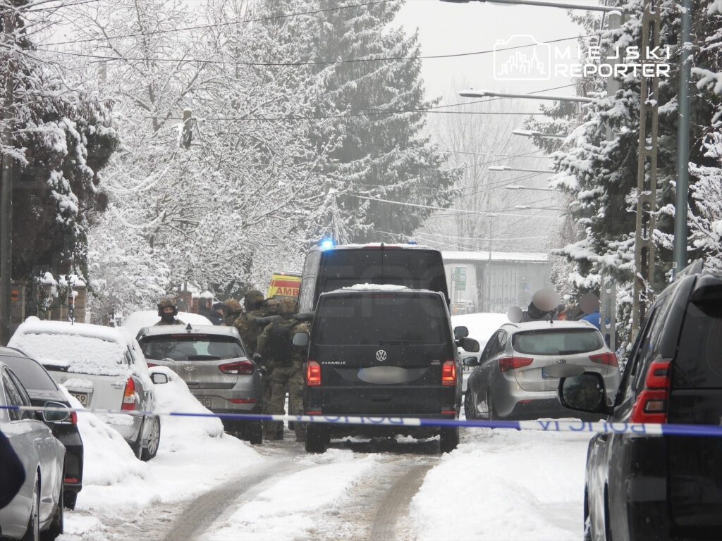 Grupa funkcjonariuszy w kamizelkach taktycznych przeszukuje teren w zaśnieżonym otoczeniu ulicy, obok zaparkowanych samochodów.