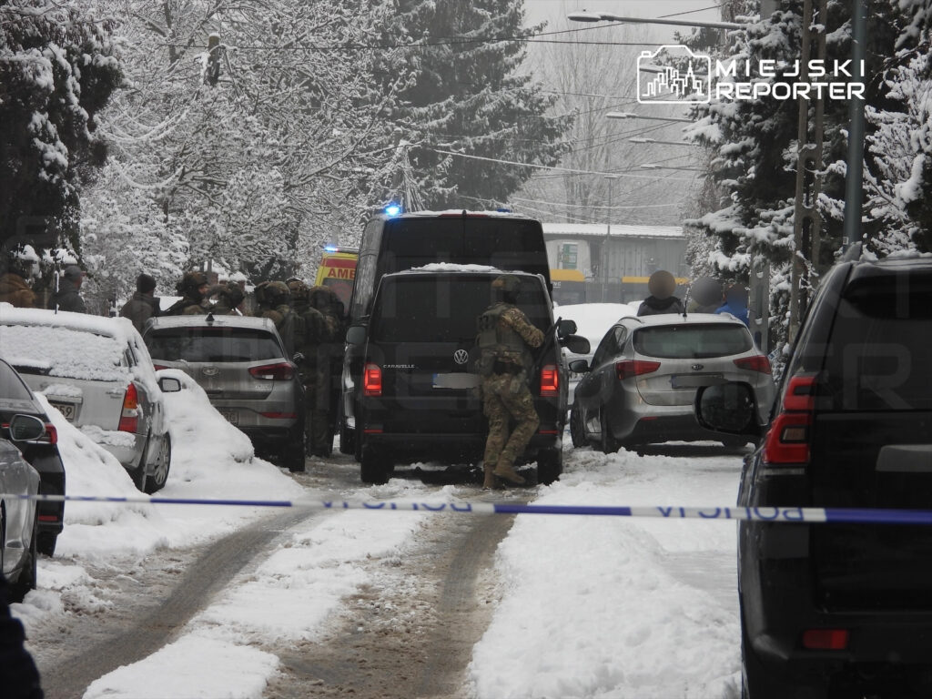 Grupa uzbrojonych mężczyzn w mundurach rozmawia obok radiowozu w zaśnieżonej ulicy, otoczonej samochodami.