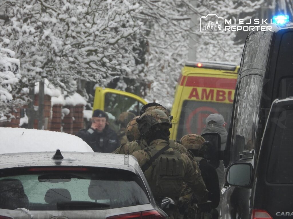 Grupa uzbrojonych funkcjonariuszy w kamizelkach taktycznych przebywa na ośnieżonej ulicy, obok ambulansu.