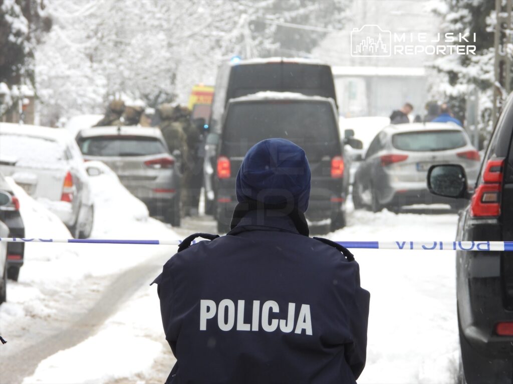 Policjant w granatowym mundurze siedzi na śniegu, obserwując działania służb w zaśnieżonej ulicy.