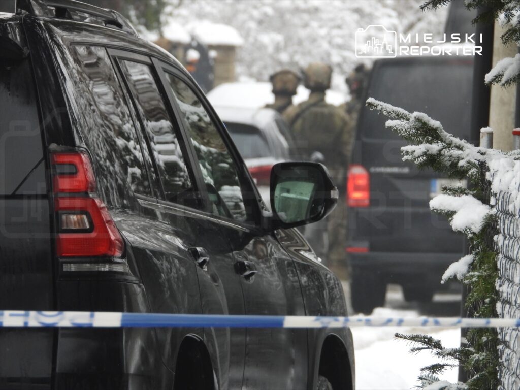 Czarny SUV zaparkowany na zaśnieżonej ulicy, w tle widoczni uzbrojeni mężczyźni w mundurach.