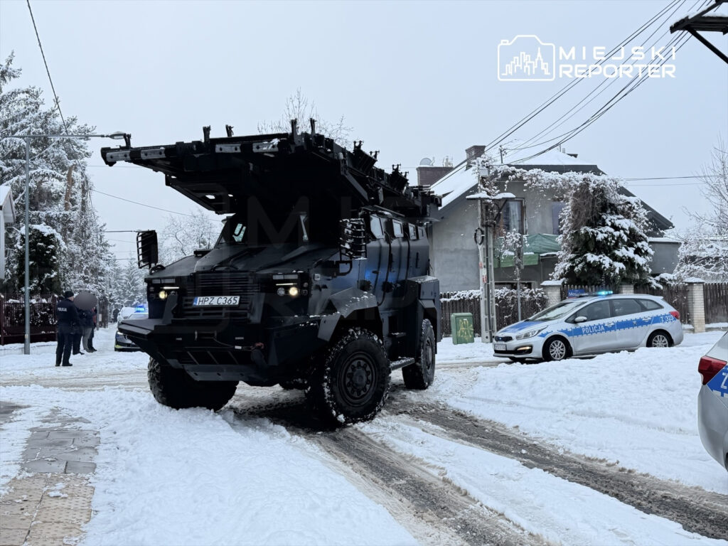 Czarny opancerzony pojazd stoi na zaśnieżonej ulicy, obok dwóch radiowozów policyjnych oraz grupy funkcjonariuszy.