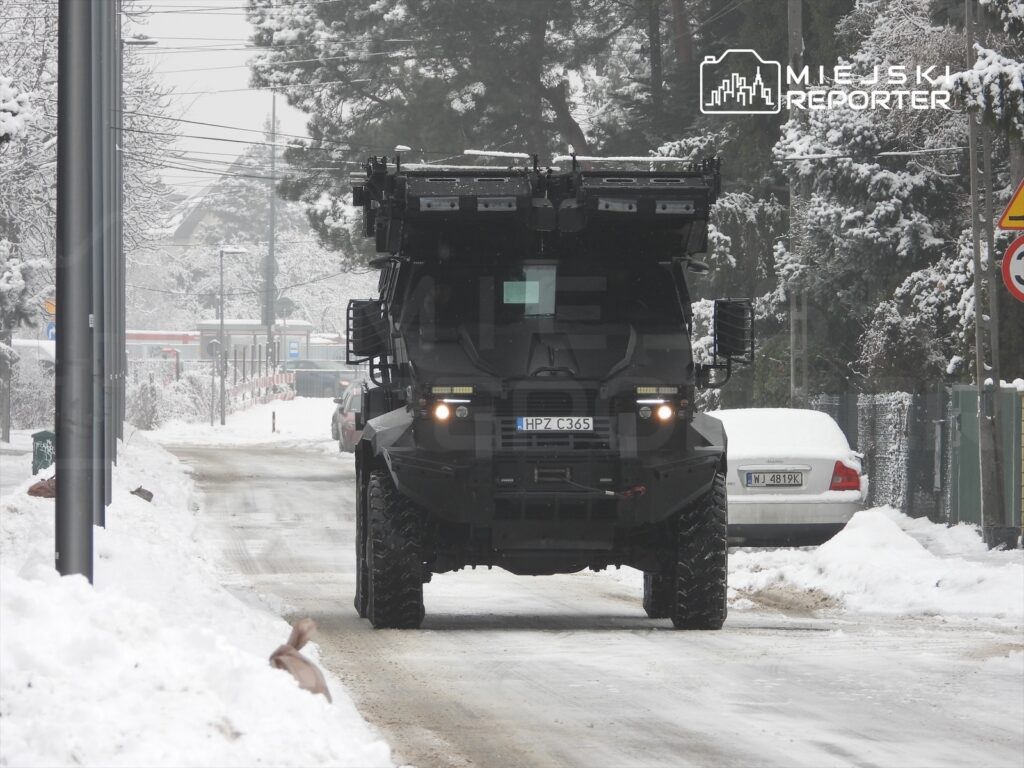 Opancerzony pojazd porusza się po zasypanej śniegiem ulicy, otoczonej drzewami i budynkami.