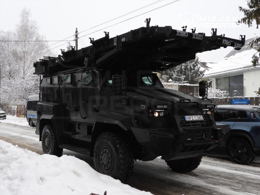 Czarny pojazd opancerzony z zamontowanym sprzętem na dachu stoi na zaśnieżonej ulicy obok innych samochodów.