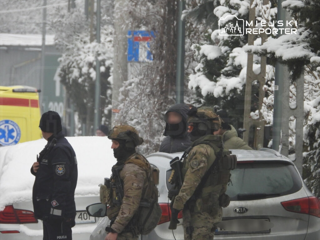 Grupa uzbrojonych funkcjonariuszy w mundurach i policjantów zabezpiecza teren w zaśnieżonej okolicy.