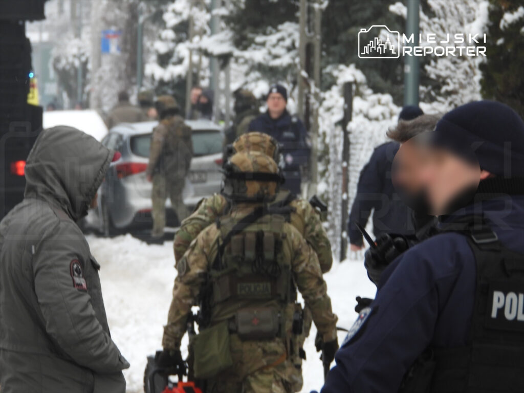 Grupa uzbrojonych żołnierzy i policjantów przeszukuje zaśnieżoną ulicę, obok stojący samochód osobowy.