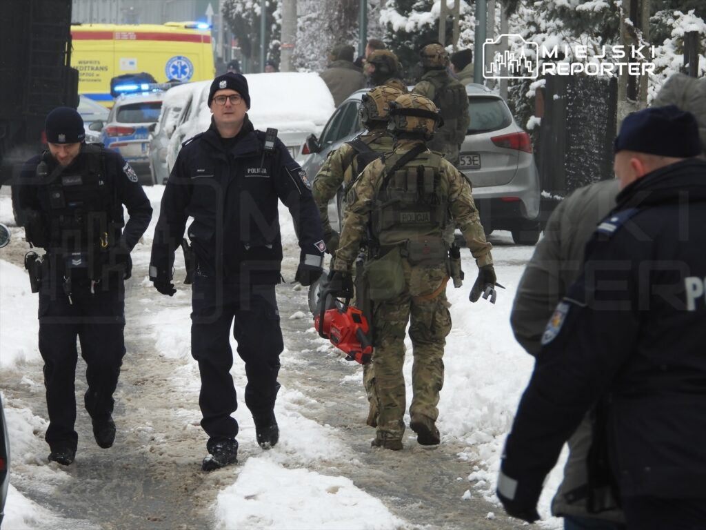 Grupa policjantów i funkcjonariuszy w umundurowaniu taktycznym porusza się po zaśnieżonej ulicy, blisko pojazdów służbowych.