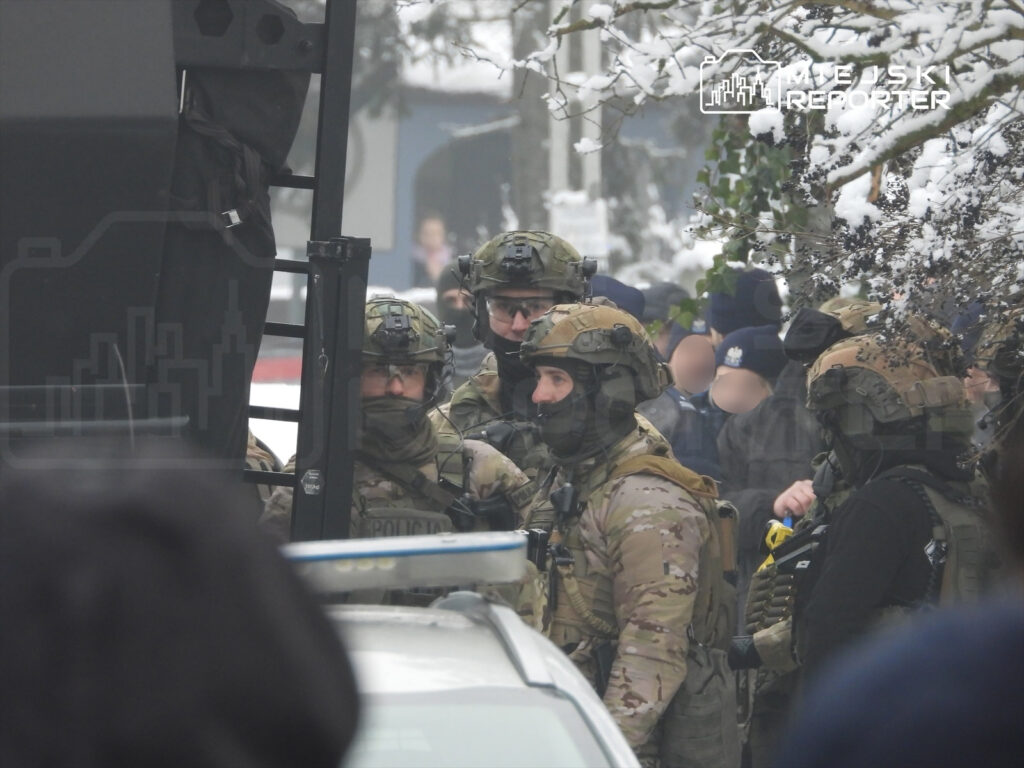 Grupa uzbrojonych funkcjonariuszy w kamizelkach taktycznych rozmawia obok pojazdu policyjnego w zaśnieżonym otoczeniu.