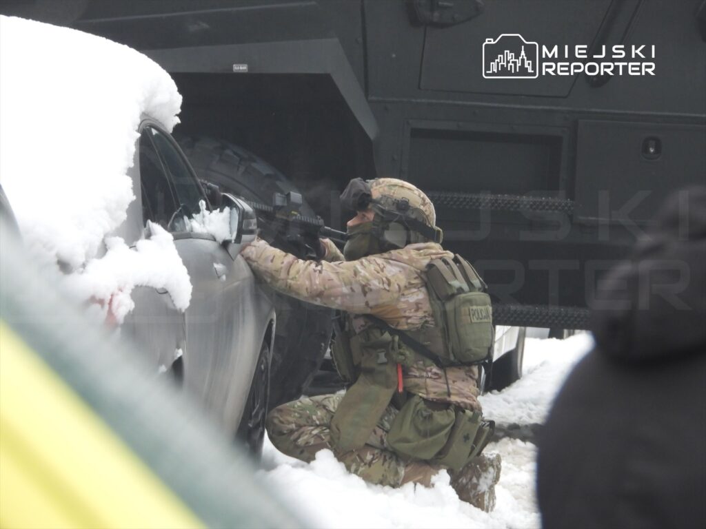 Żołnierz w mundurze kamuflażowym przyjmuje pozycję za samochodem, trzymając broń w kierunku nieznanego celu. Wokół leży śnieg.