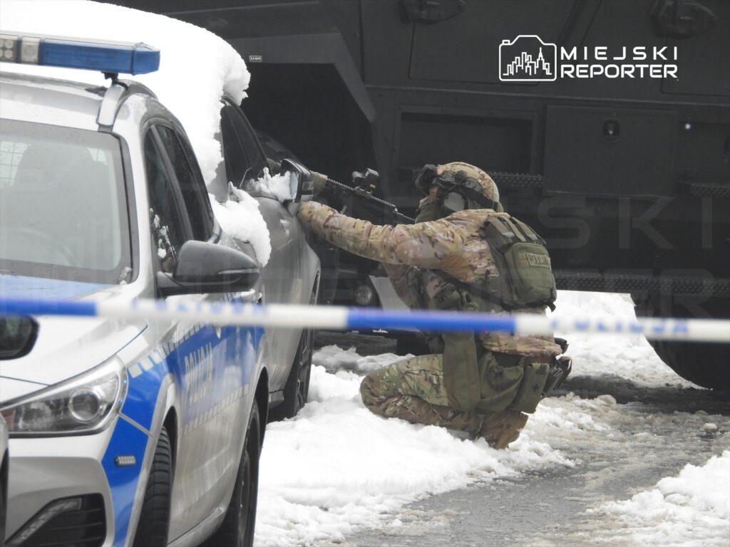 Żołnierz w kamizelce taktycznej klęczy obok samochodu pokrytego śniegiem, trzymając broń w kierunku pojazdu.