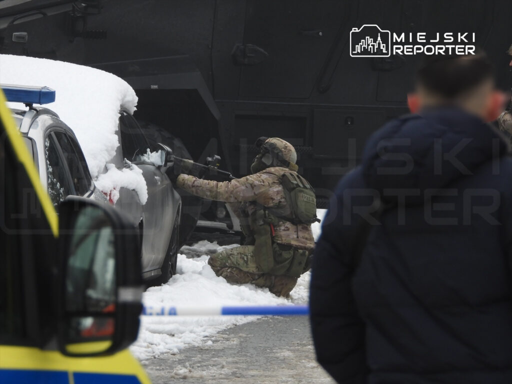 Żołnierz w kamuflażu klęczy przy samochodzie pokrytym śniegiem, trzymając broń w kierunku pojazdu.