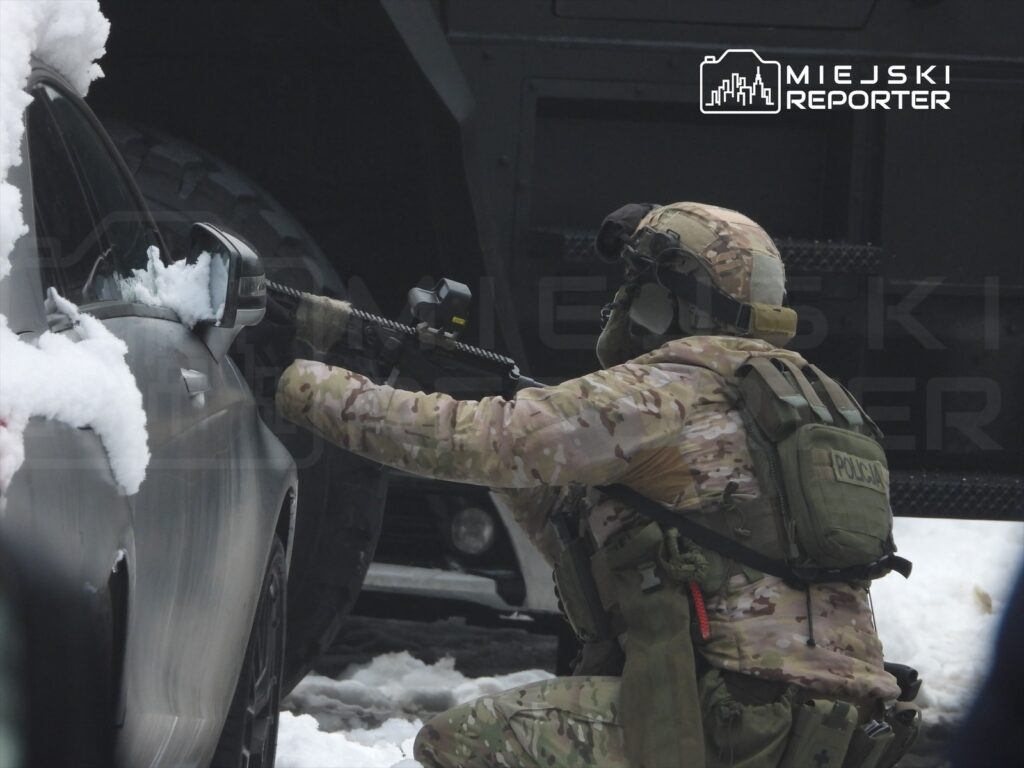 Żołnierz w kamuflażu, trzymając broń, przeszukuje samochód pokryty śniegiem na zaśnieżonej ulicy.