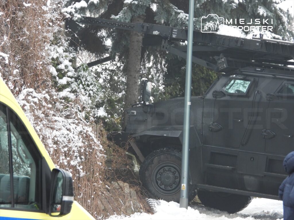 Czarny pojazd opancerzony stoi na śniegu obok zarośli, a w tle widać niebieski ambulans.
