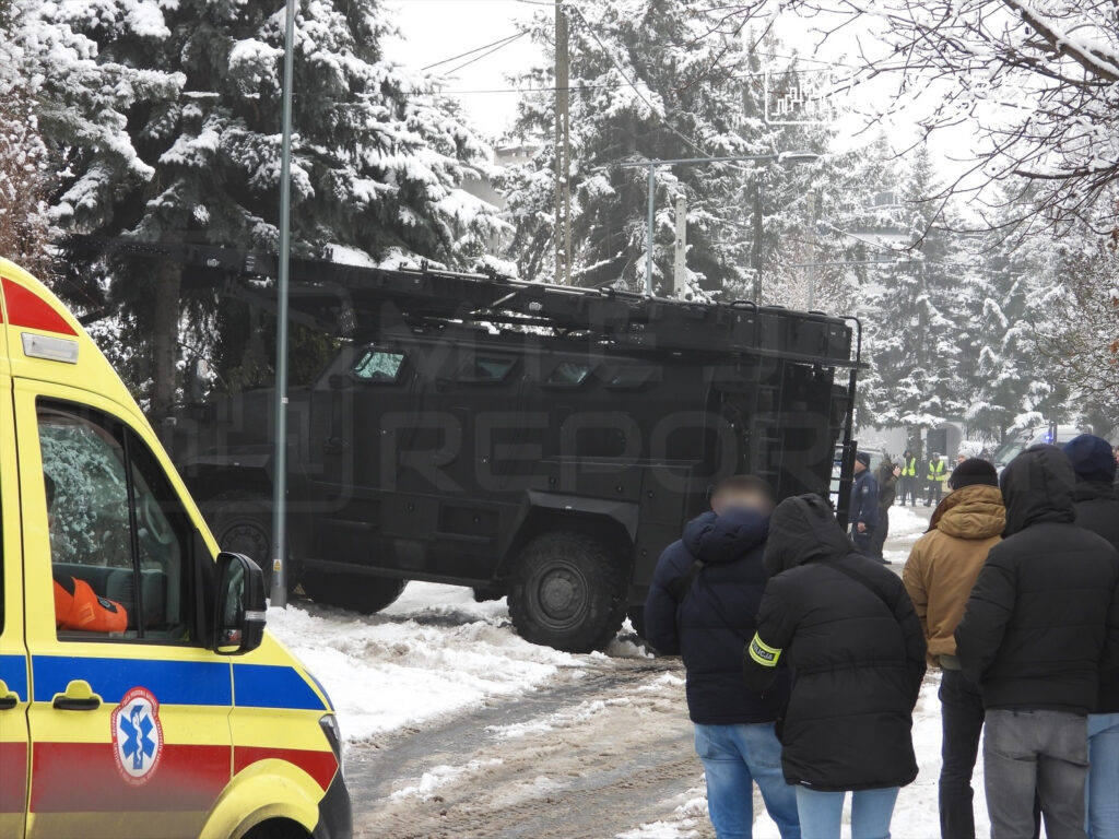 Grupa osób w zimowych kurtkach obserwuje policyjny pojazd opancerzony na zaśnieżonej ulicy. W tle widoczne drzewa pokryte śniegiem.