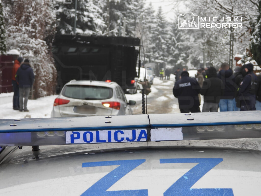 Grupa policjantów w mundurach rozmawia na zaśnieżonej ulicy, obok zaparkowanego samochodu.