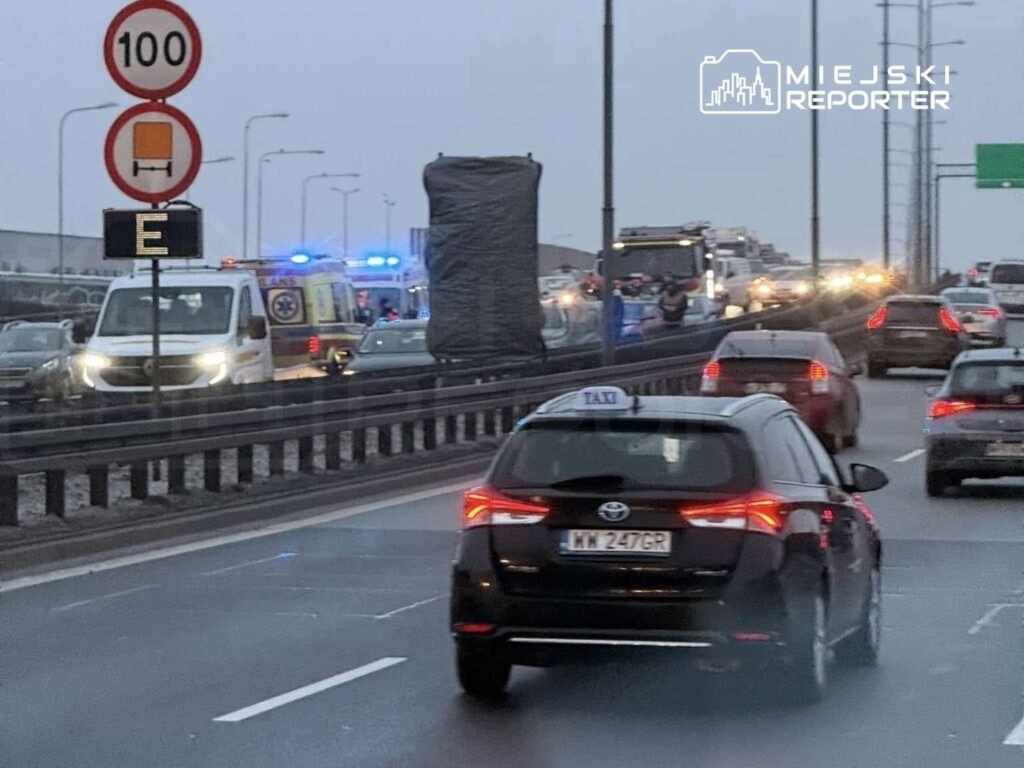 Kierowca taksówki porusza się w korku na drodze, obok widoczna karetka pogotowia oraz pojazdy policyjne.