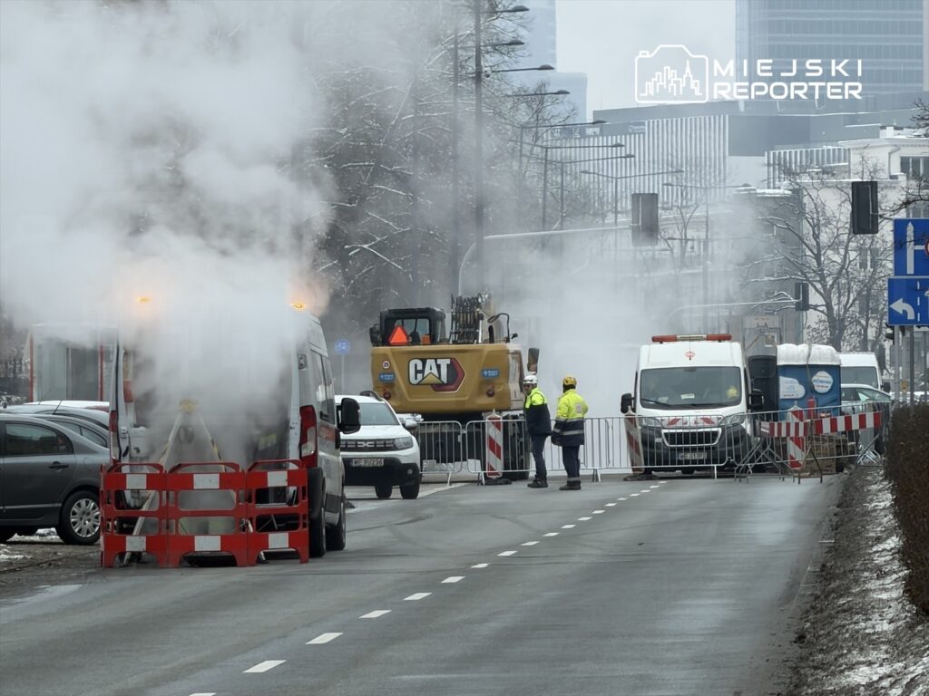 Na ulicy unosi się para, a pracownicy w odblaskowych kamizelkach monitorują prace budowlane przy zamkniętej drodze.