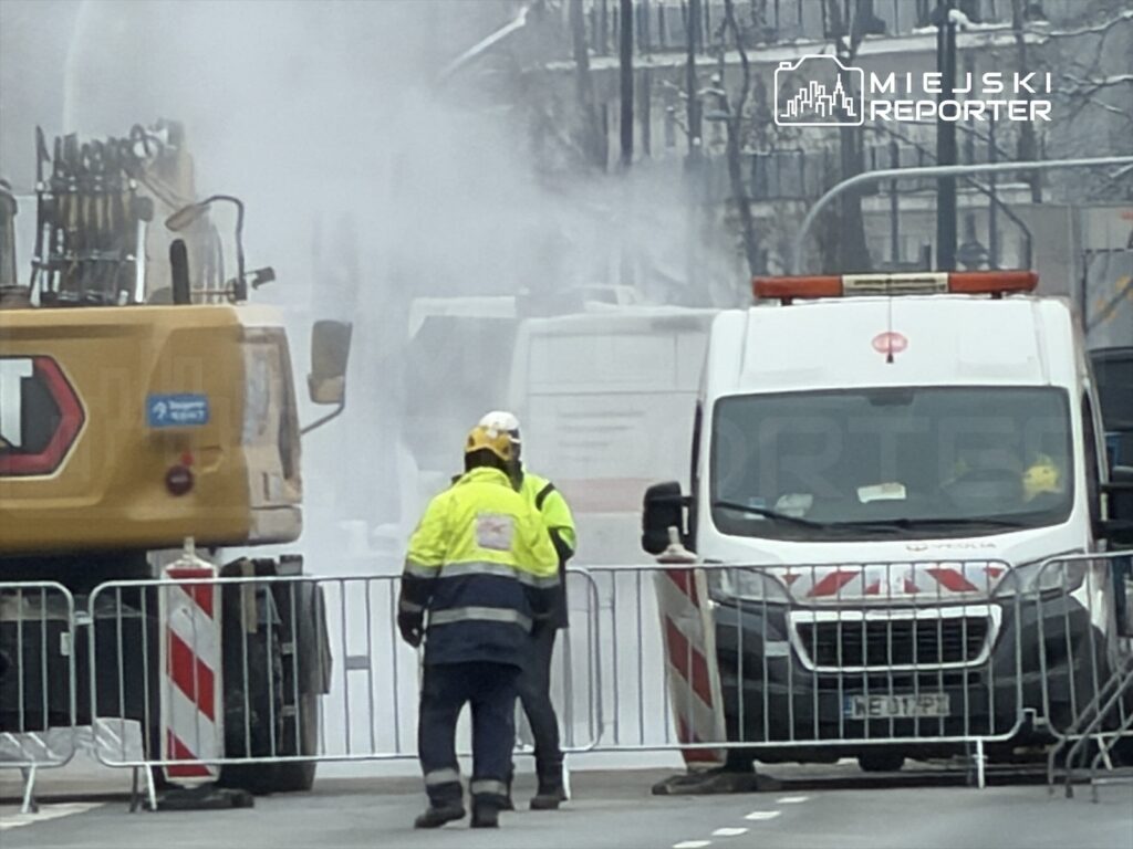 Robotnicy w odblaskowych kurtkach oceniają sytuację w rejonie wycieku pary wodnej na ulicy, obok sprzętu budowlanego.