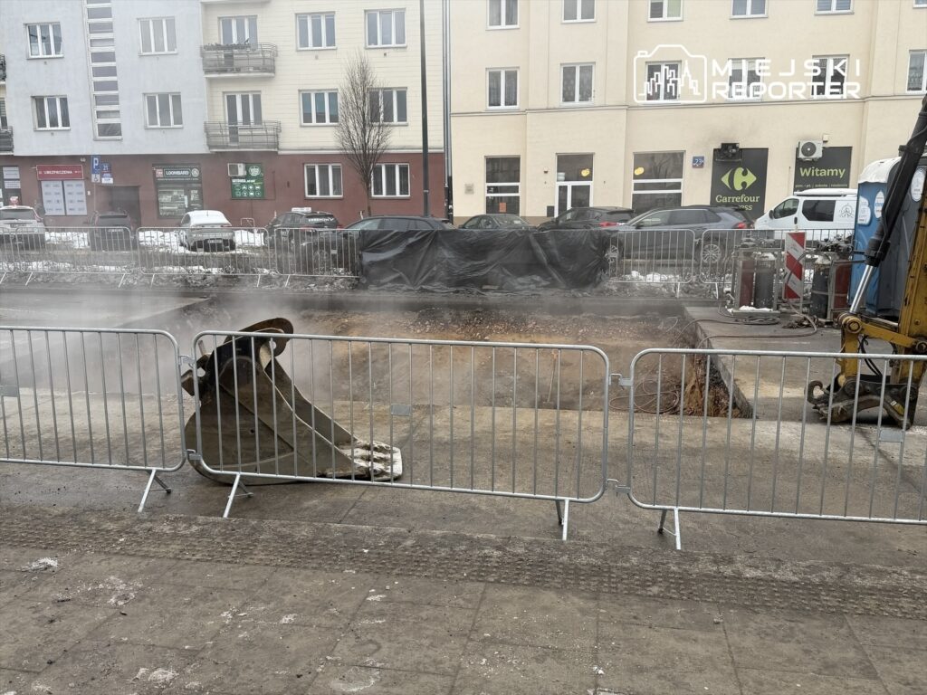 Wykop budowlany z widoczną łyżką koparki, otoczony barierkami, z parą unoszącą się nad ziemią.