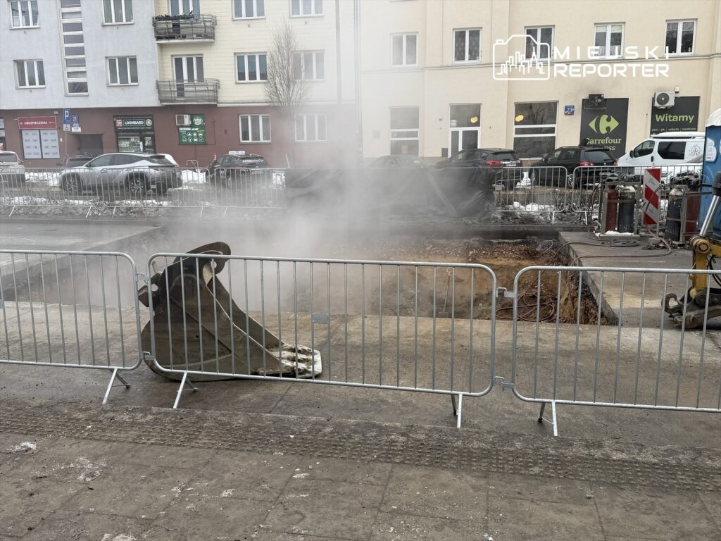 W miejscu budowy unosi się para z wykopu, obok widoczna koparka i metalowe ogrodzenie.