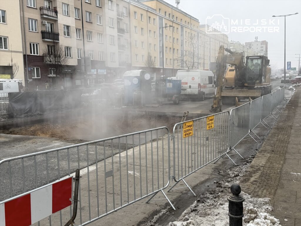 Maszyna budowlana wykonująca wykop na ulicy, otoczona barierkami ochronnymi, w tle bloki mieszkalne i pojazdy.