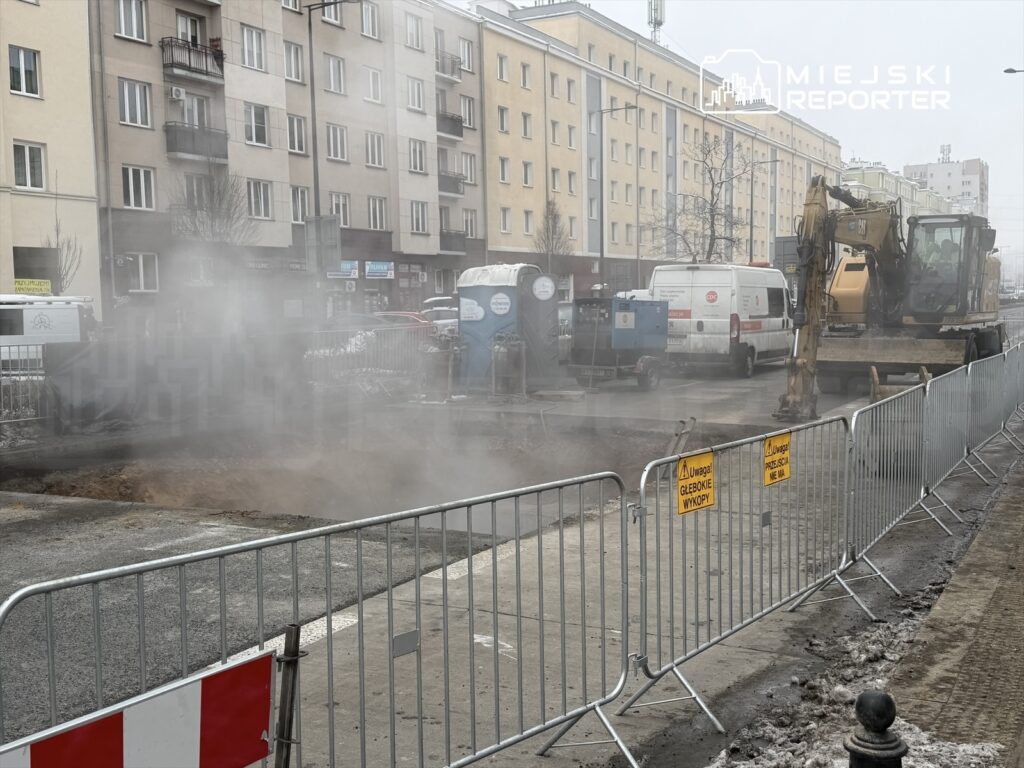 Maszyna budowlana wykonująca wykopy na ulicy, otoczona barierkami i unoszącym się pyłem. W tle widoczne bloki mieszkalne.