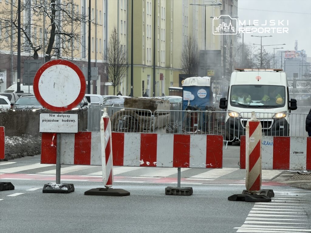 Robotnicy przy pracy w strefie budowy, z czerwono-białymi barierkami i znakiem zakazu wjazdu dla pojazdów.