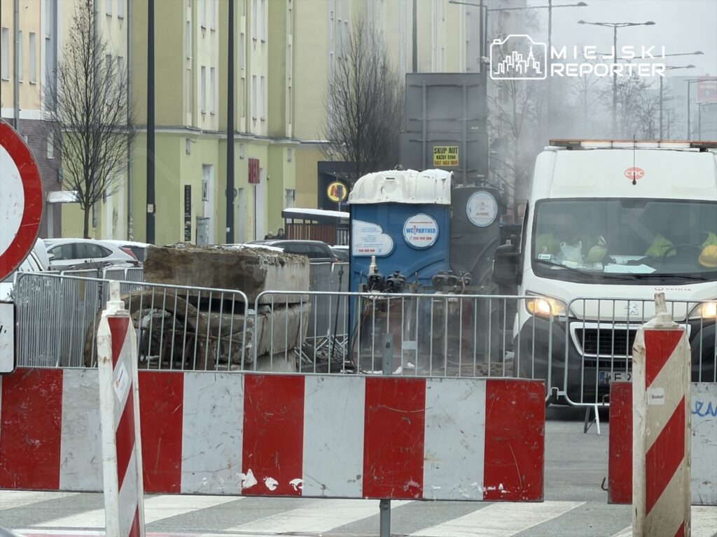 Robotnicy prowadzą prace budowlane w zatłoczonej ulicy, otoczeni barierkami i znakami drogowymi.