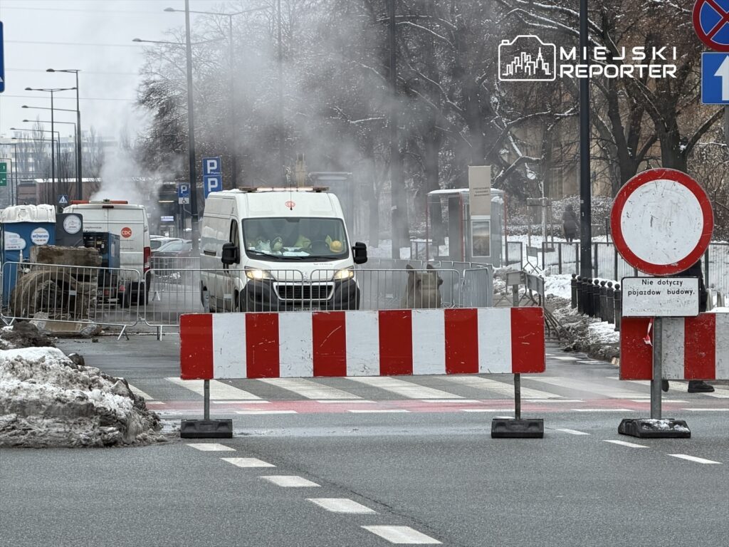 Na drodze zamkniętej dla ruchu stoją samochody dostawcze, a w tle unosi się para z wyrobisk budowlanych.