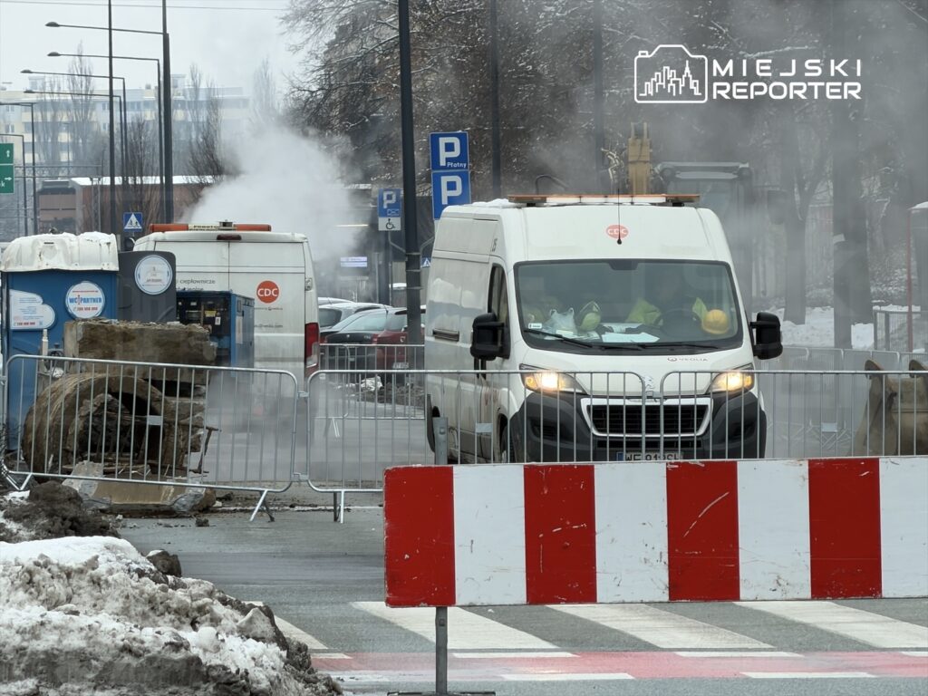 Biały furgon z logo CDC przejeżdża przez teren budowy, gdzie widać parę i barierki ochronne.