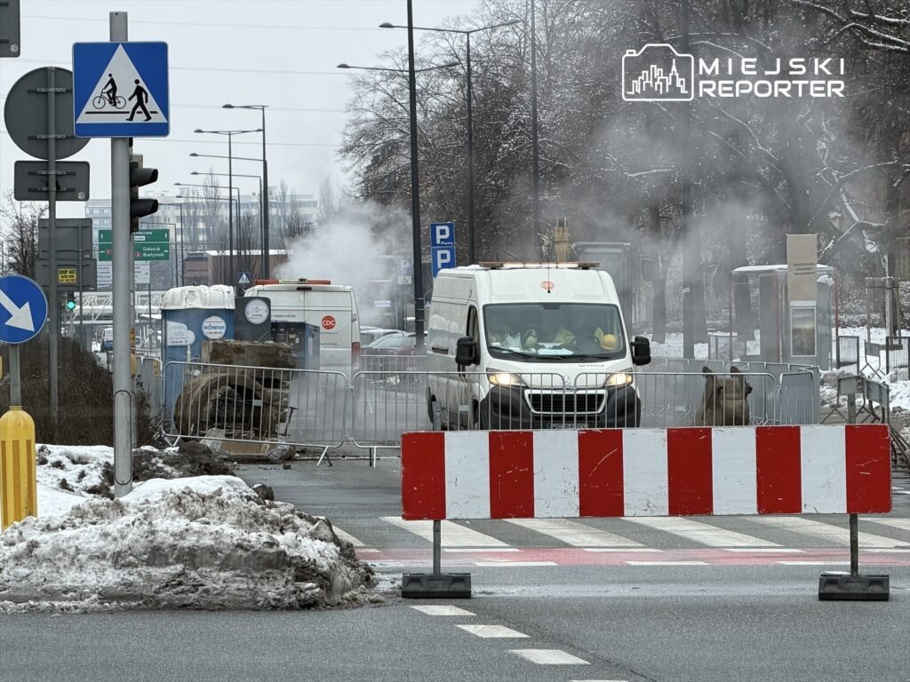 Biały furgon przejeżdża przez zablokowaną ulicę, w tle widoczne prace budowlane i unosząca się para.
