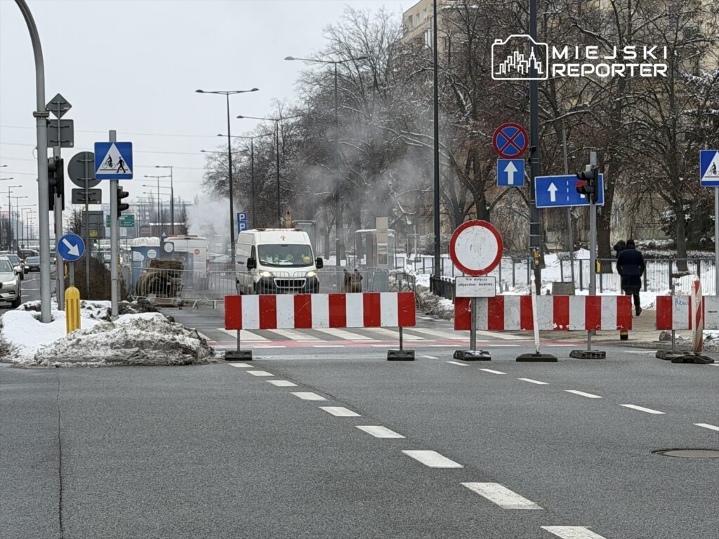 Biały furgon przejeżdża obok barier ustawionych na ruchliwej ulicy, w tle widoczne unoszące się opary.