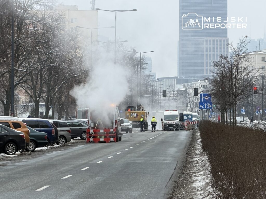 Ekipa budowlana pracująca na ulicy, obok unoszącego się dymu z pojazdu, w tle nowoczesne budynki.