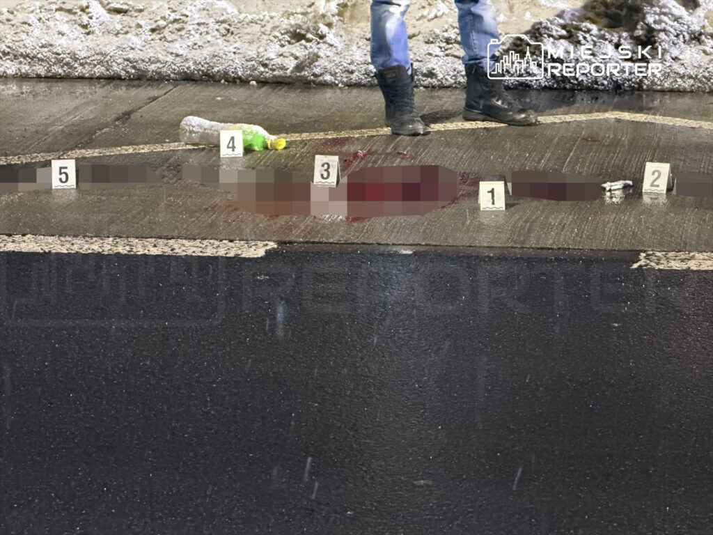 Na jezdni leży ciało, wokół oznaczenia dowodowe oraz butelka, a w tle widać śnieg.