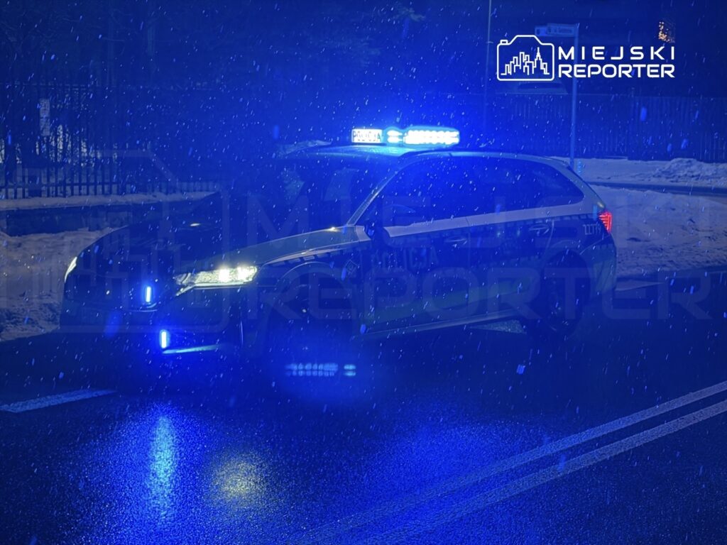 Policyjny radiowóz z włączonymi niebieskimi światłami stoi na oświetlonej ulicy w trakcie opadu śniegu.