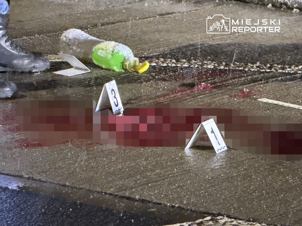 Mężczyzna w butach roboczych stoi obok oznaczeń na jezdni, gdzie znajduje się plama krwi i butelka.