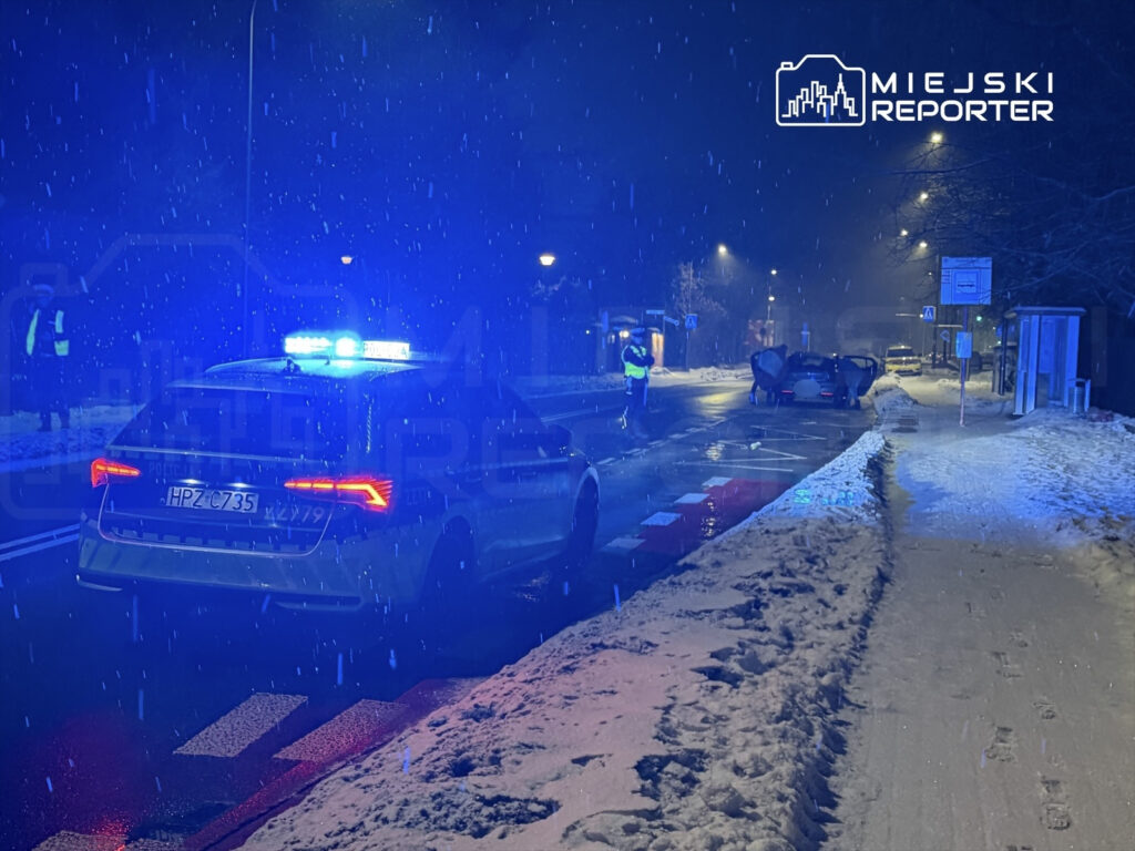 Policyjny radiowóz z włączonymi sygnałami świetlnymi stoi na ośnieżonej ulicy, obok rozbitego samochodu.