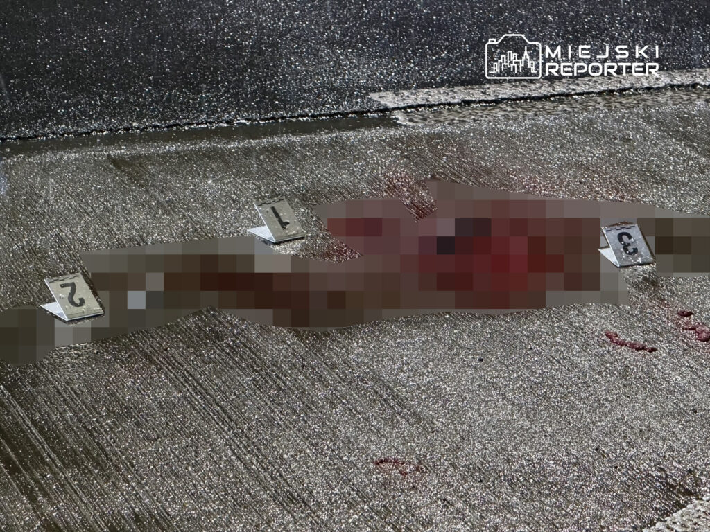 Na ulicy leży zakrwawiona osoba, obok której znajdują się oznaczenia dowodowe.
