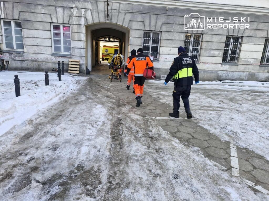 Ratownicy medyczni i strażnik miejski wchodzą do budynku, niosąc nosze w otoczeniu śnieżnej nawierzchni.