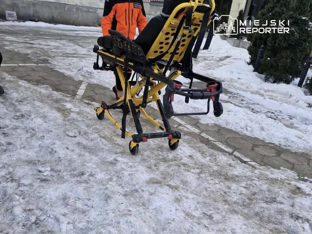 Ratownik medyczny przemieszcza nosze w zimowym otoczeniu, na pokrytej śniegiem ulicy.