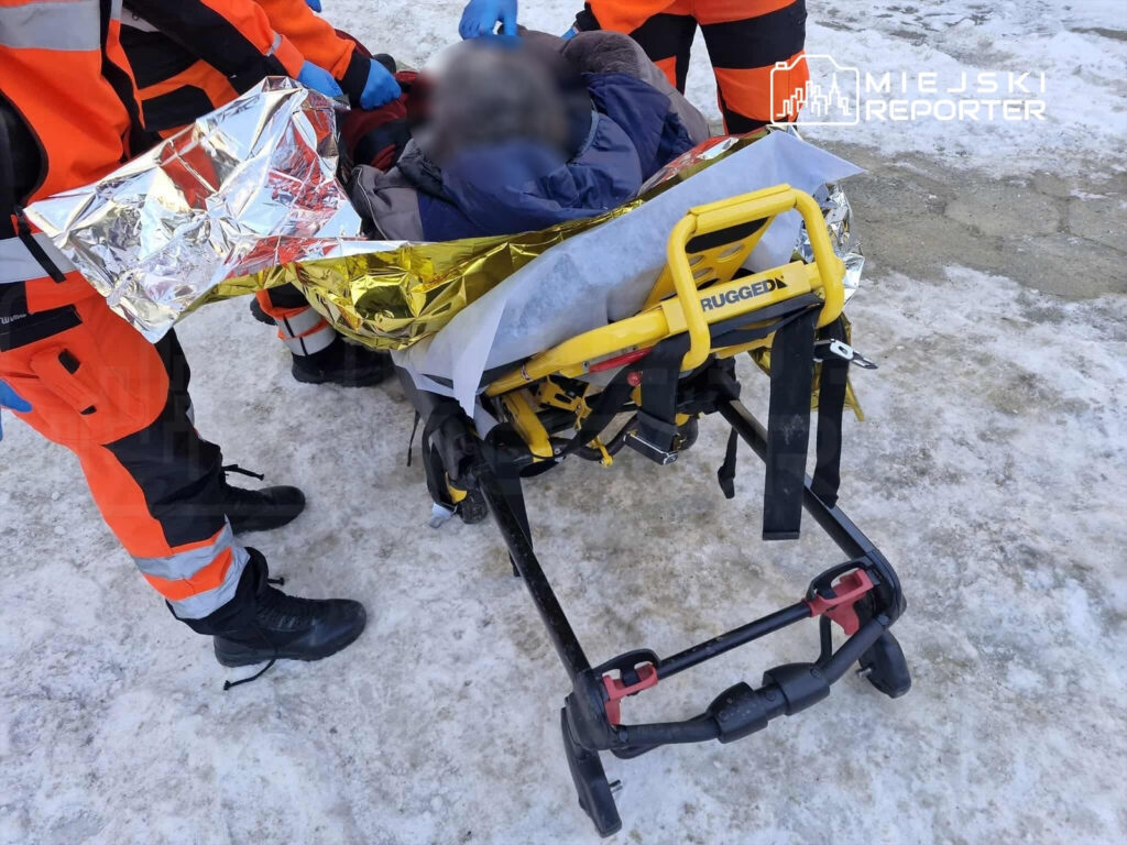 Ratownicy medyczni przenoszą osobę na noszach z folią termiczną w zimowym otoczeniu.