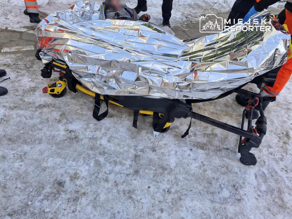 Ratownicy medyczni zabezpieczają osobę na noszach pokrytych folią termiczną na zaśnieżonym terenie.