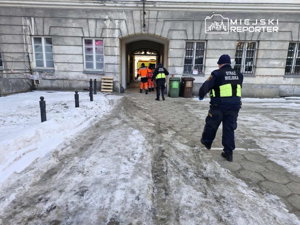Grupa funkcjonariuszy Straży Miejskiej w mundurach przemieszcza się w kierunku wejścia do budynku, obok śnieżnej nawierzchni.