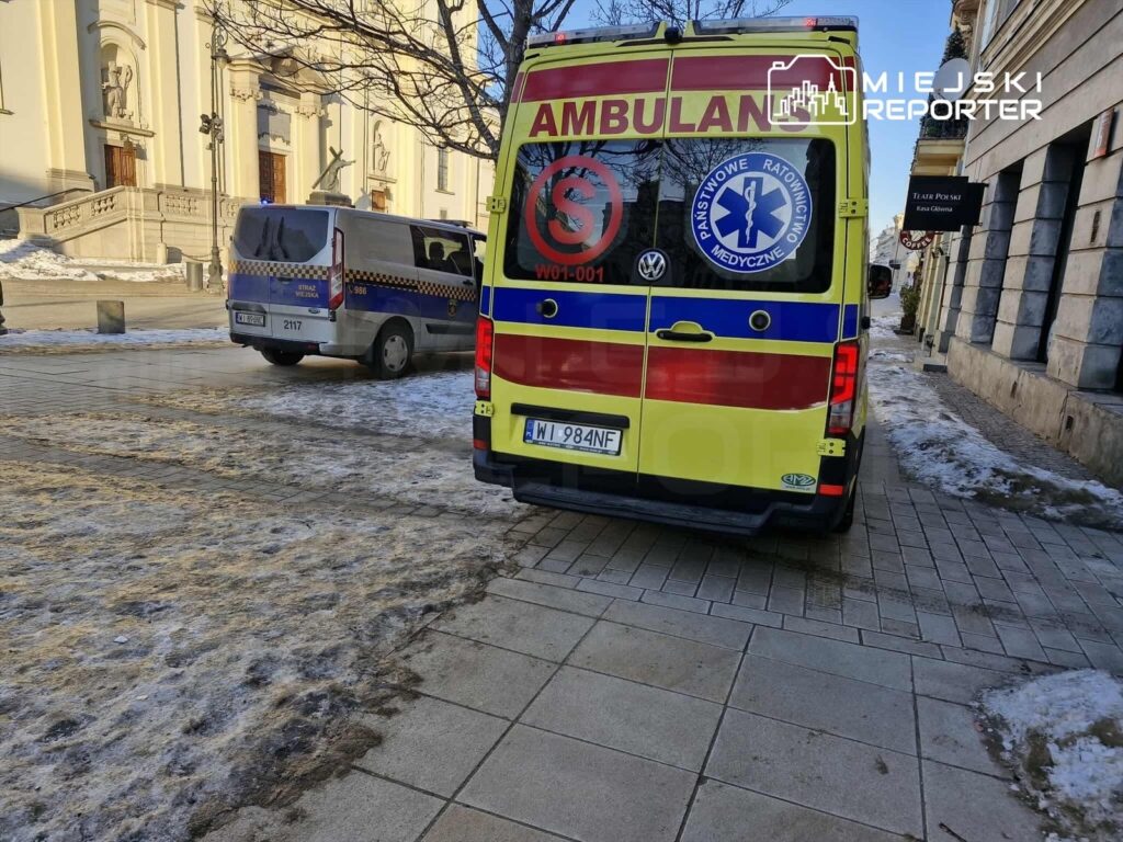 Ambulans z włączonymi sygnałami świetlnymi stoi na chodniku obok budynku z klasyczną architekturą.