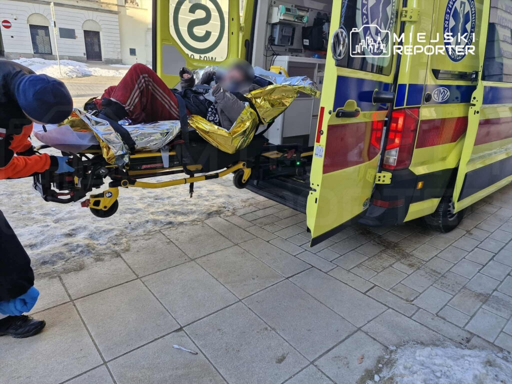 Ratownik medyczny przystosowuje nosze z pacjentem w kocach termicznych do transportu do karetki na zaśnieżonej ulicy.