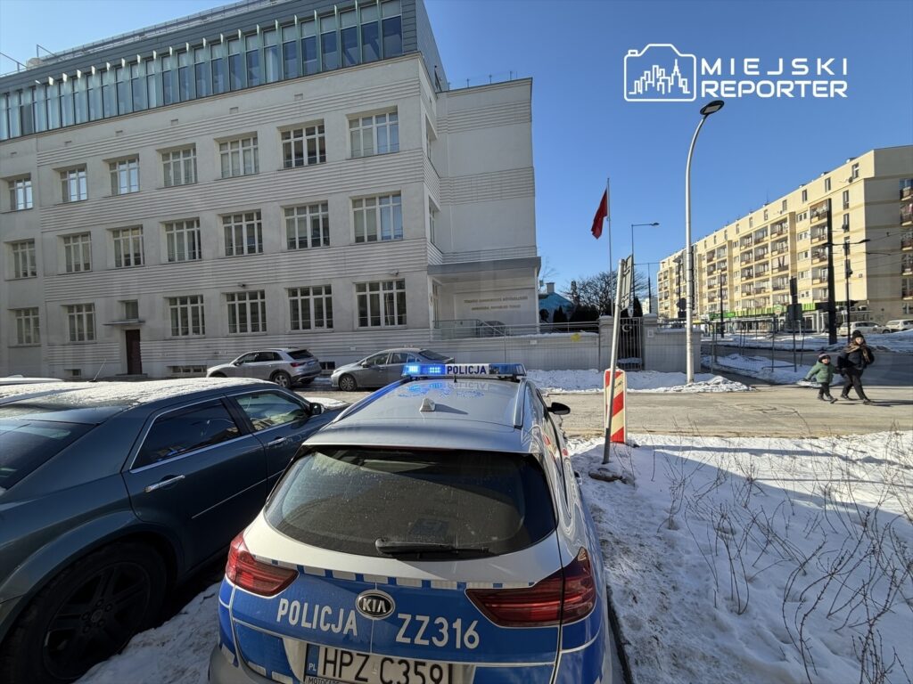 Policyjny radiowóz z włączonymi sygnalizatorami świetlnymi stoi na zaśnieżonej ulicy obok nowoczesnego budynku.