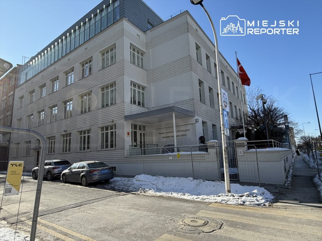 Nowoczesny budynek z przeszkloną fasadą, przed którym stoją dwa samochody, z flagą Turcji na maszcie.