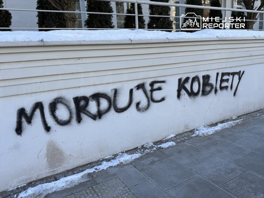 Na białej ścianie budynku widoczny jest czarny spray z napisem "MORDUJE KOBIETY". Obok leży śnieg.