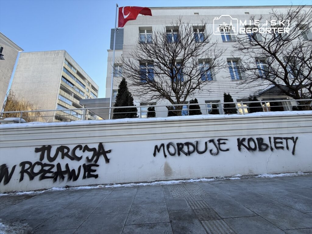 Na białej ścianie budynku widoczne są napisy sprayem: "TURCJA W ROZWAIE" oraz "MORDUJE KOBIETY". W tle widać flagę Turcji.