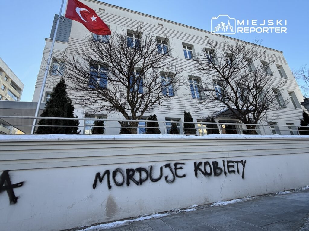 Biały budynek z flagą Turcji, na ścianie widoczny napis "MORDUJE KOBIETY" w czarnym sprayu.